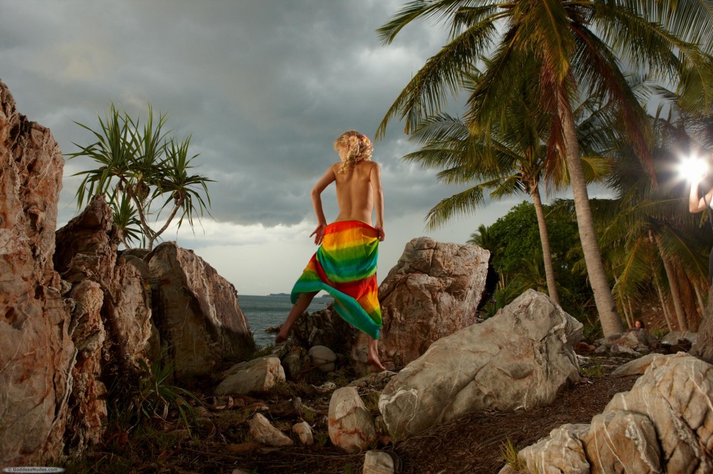 Model swishes bright fabric around her nude and sleek body on tropical rocky beach